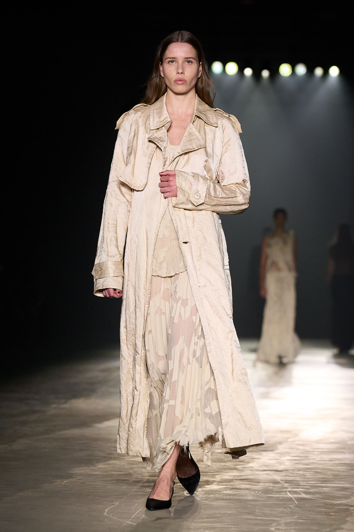 Model walking a fashion runway in a long beige trench over a pale patterned dress, black heels, under spotlights.