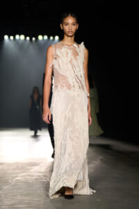Female model walking a fashion runway in a sleeveless, cream-toned, textured gown with layered fabric details under bright runway lights.
