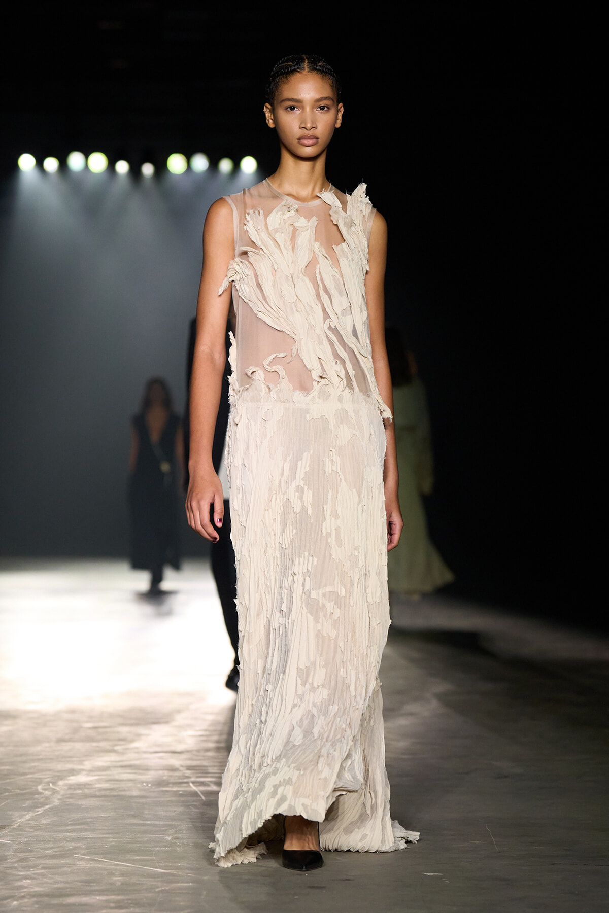 Female model walking a fashion runway in a sleeveless, cream-toned, textured gown with layered fabric details under bright runway lights.