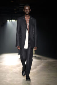 Male model on a runway wearing a loose black blazer with white piping, black pants, and black boots.