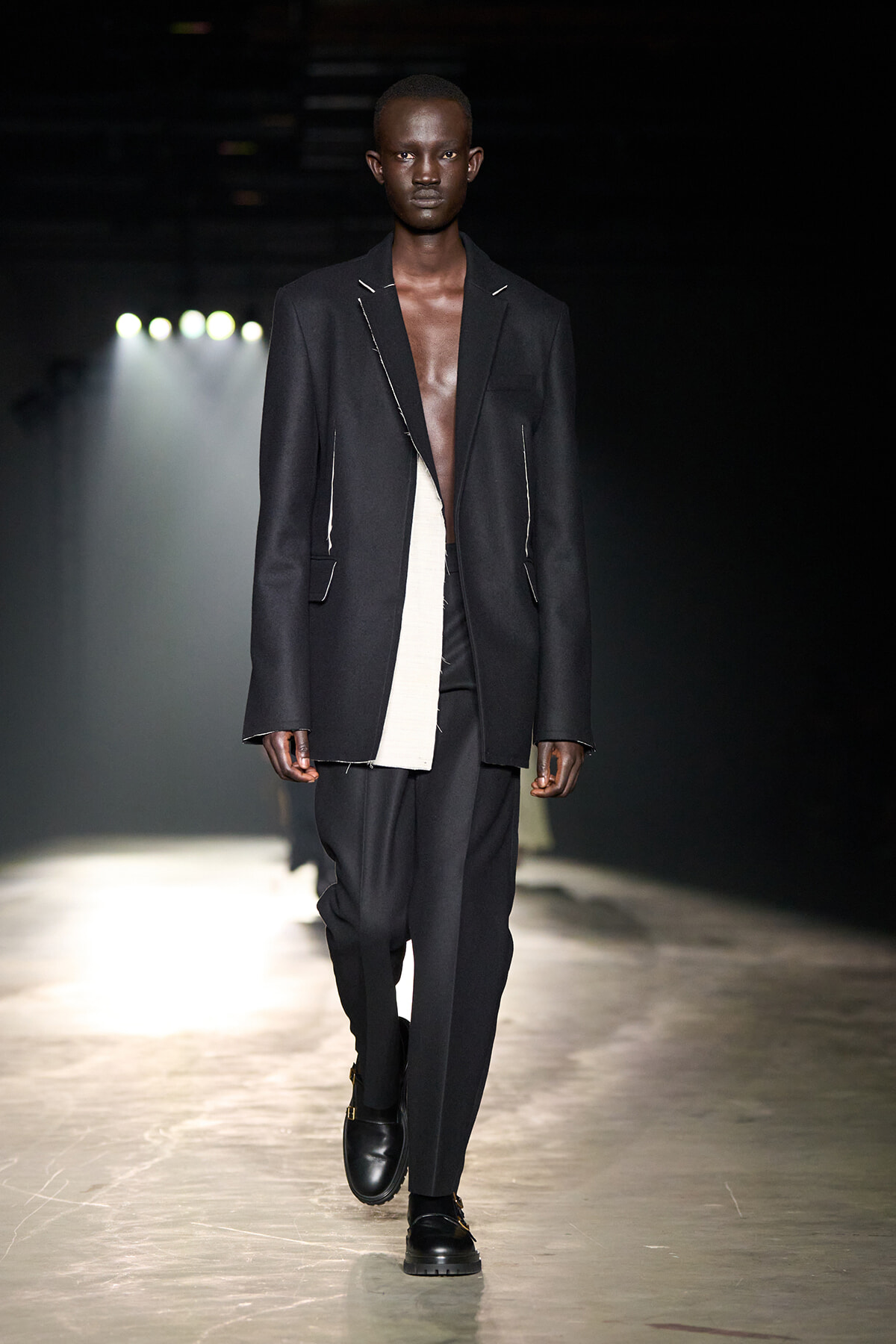 Male model on a runway wearing a loose black blazer with white piping, black pants, and black boots.