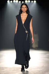 Model walking a runway in a long black sleeveless dress with a deep V neckline and a wrap detail on the front.