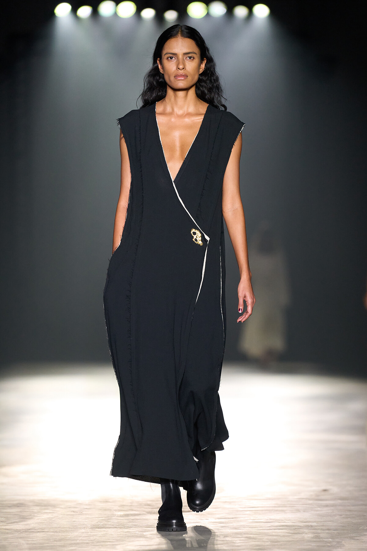 Model walking a runway in a long black sleeveless dress with a deep V neckline and a wrap detail on the front.