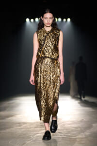 Model walks down a runway wearing a sleeveless metallic gold textured dress with a black belt, black footwear, under strong spotlight.