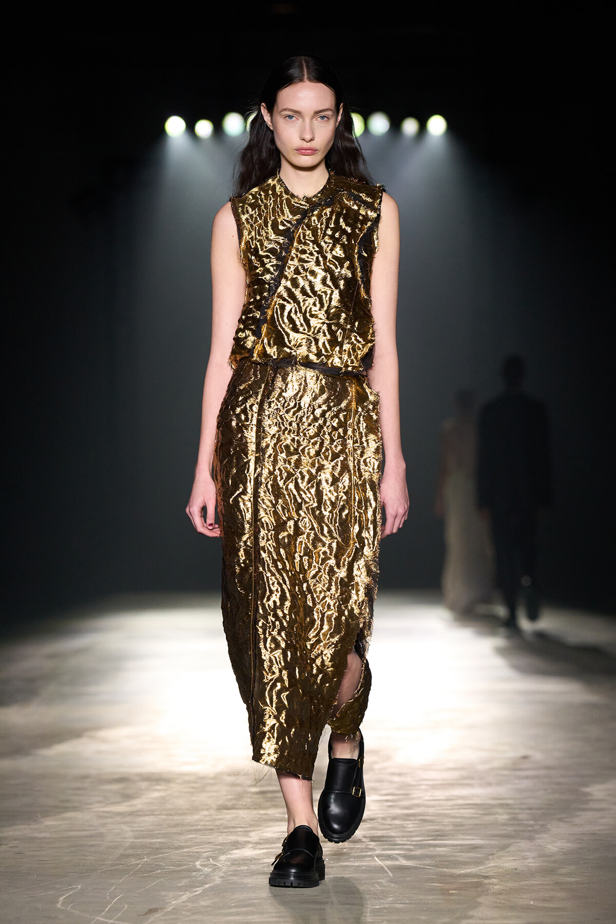 Model walks down a runway wearing a sleeveless metallic gold textured dress with a black belt, black footwear, under strong spotlight.