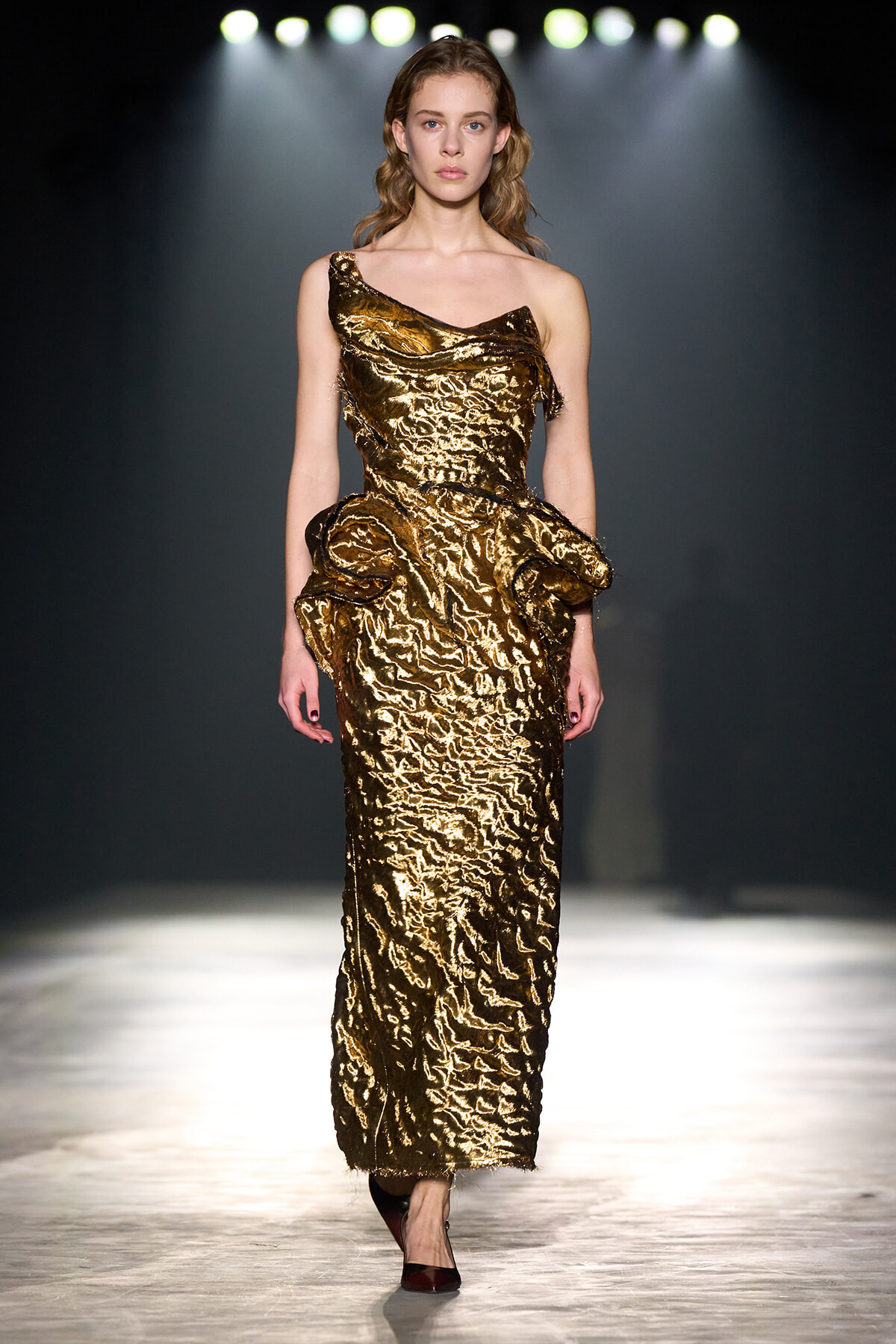 Model walks the runway in a metallic gold one-shoulder dress with sculpted ruffles.