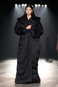 Model walks the runway wearing a long, oversized black coat under stage lights on a fashion show.