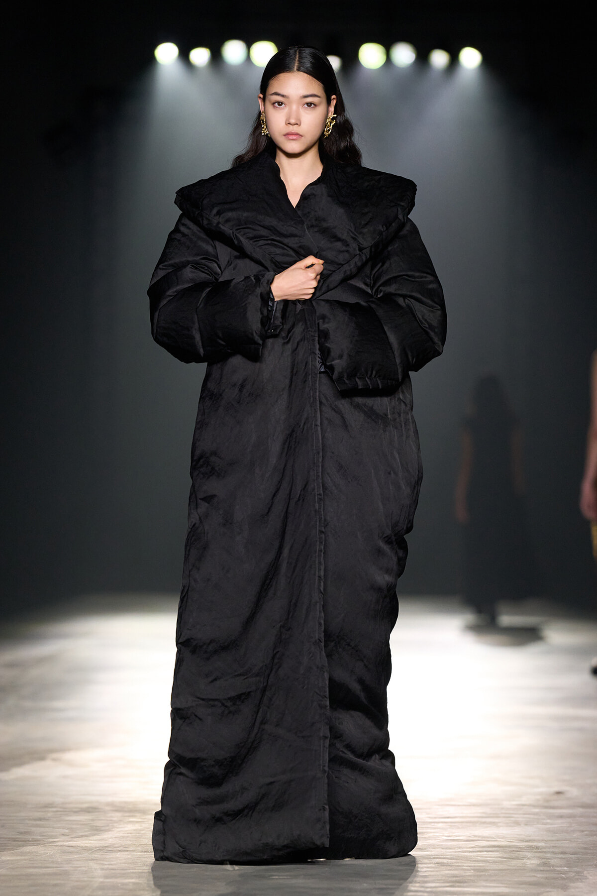 Model walks the runway wearing a long, oversized black coat under stage lights on a fashion show.