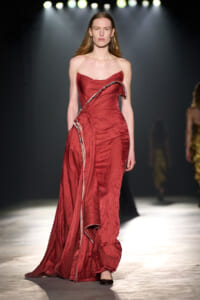 Model walking a fashion runway in a strapless, crimson red gown with draped, ruffled detailing and a long trailing train under spotlight.
