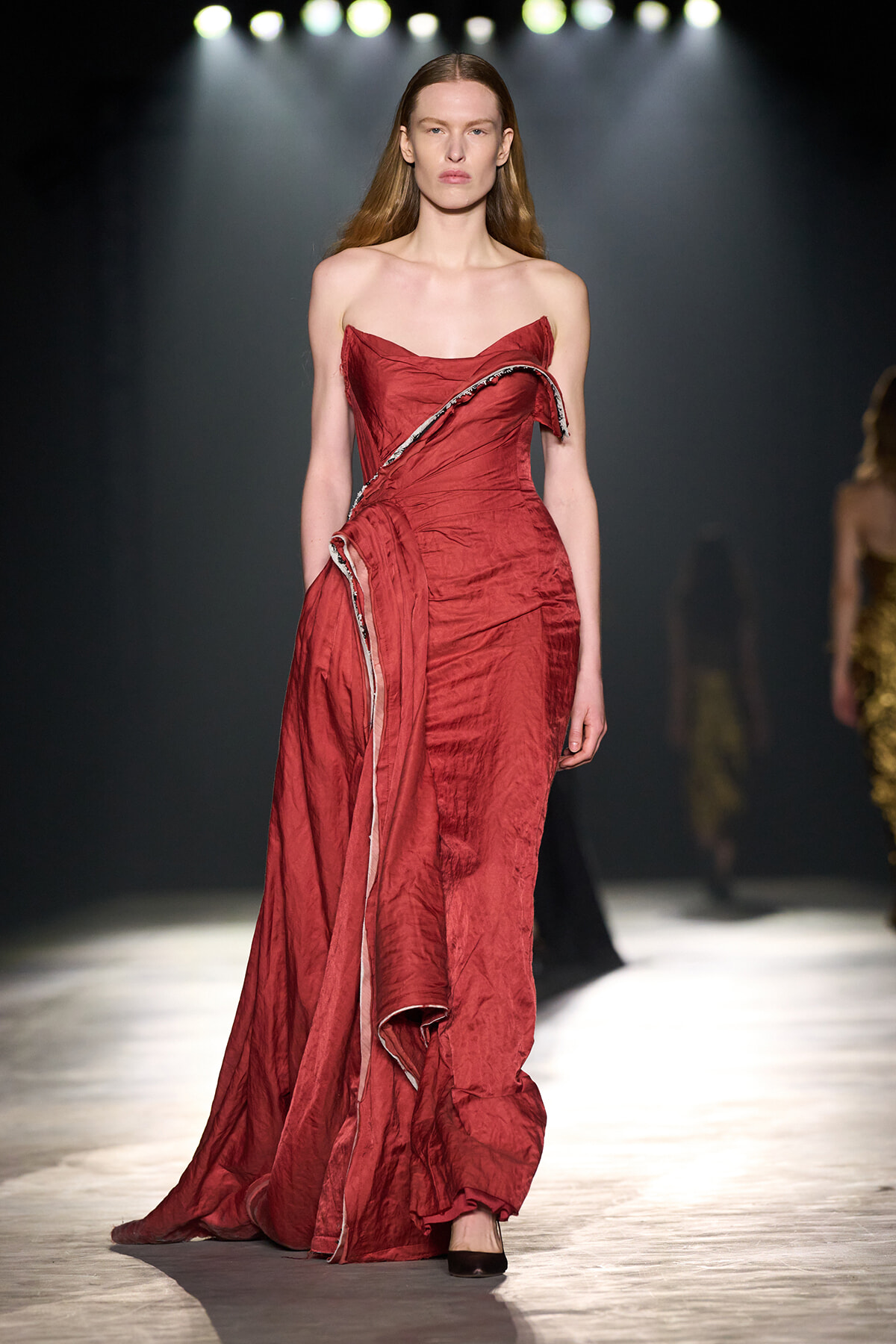 Model walking a fashion runway in a strapless, crimson red gown with draped, ruffled detailing and a long trailing train under spotlight.