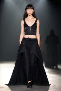 Model walking a runway in a black two-piece ensemble: cropped top with seam details and a long, layered asymmetric skirt with white stitching, under studio lights.