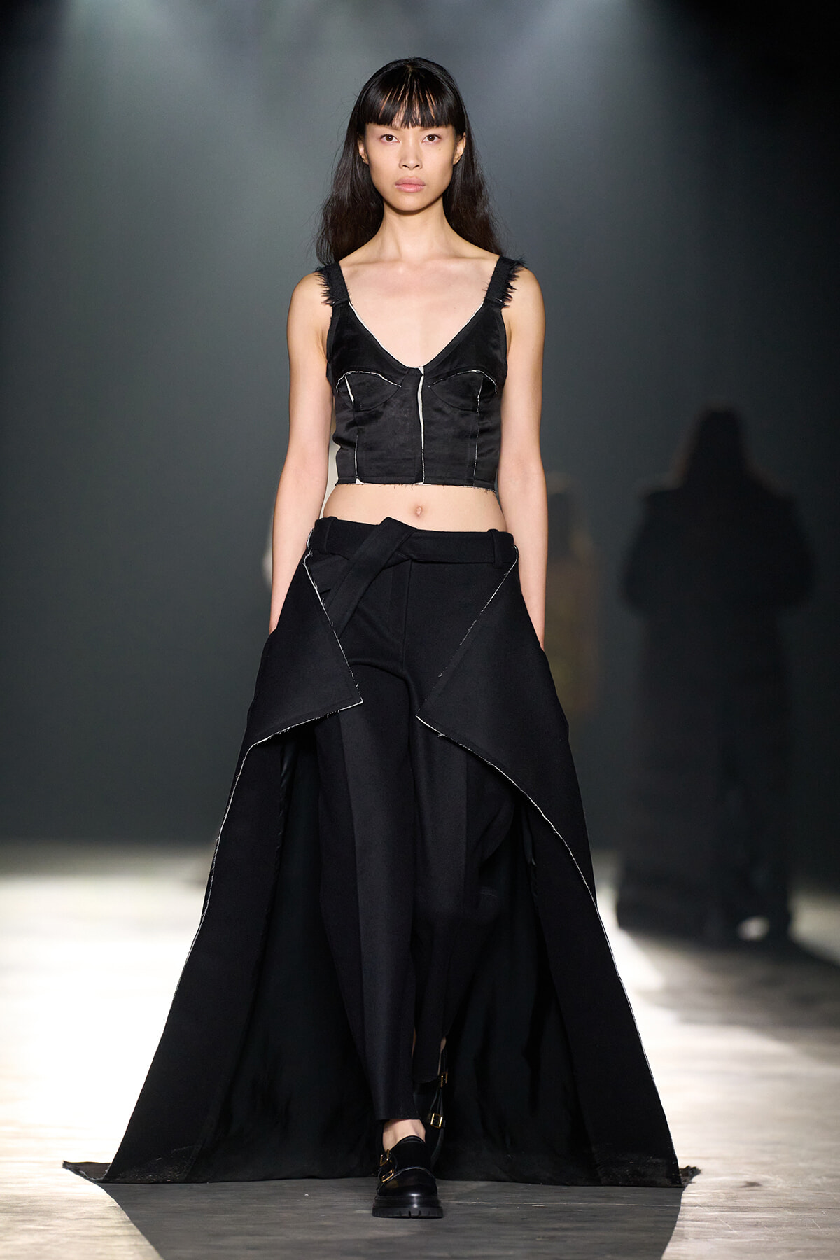 Model walking a runway in a black two-piece ensemble: cropped top with seam details and a long, layered asymmetric skirt with white stitching, under studio lights.
