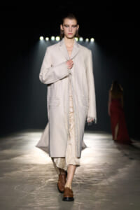 Model walks the runway in a cream long coat with frayed edges and brown loafers under spotlights.