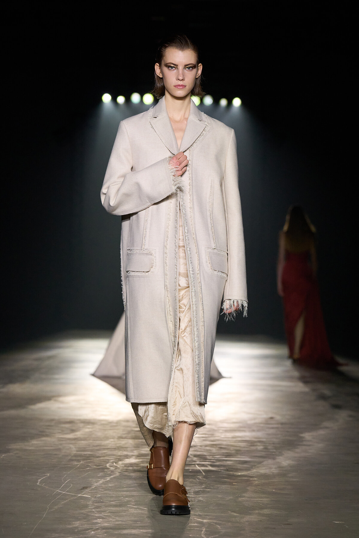 Model walks the runway in a cream long coat with frayed edges and brown loafers under spotlights.