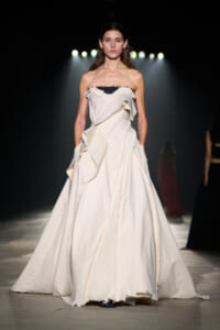 Model on a dark runway wearing a strapless ivory gown with large cascading ruffles and a raw hem on the skirt.