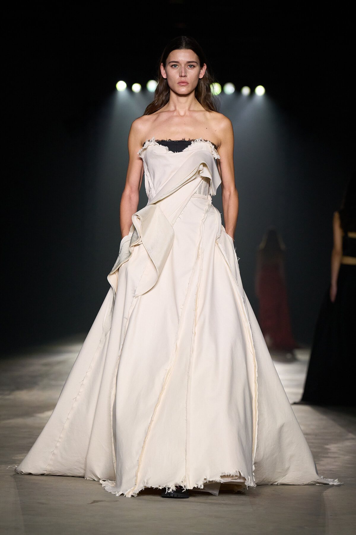 Model on a dark runway wearing a strapless ivory gown with large cascading ruffles and a raw hem on the skirt.