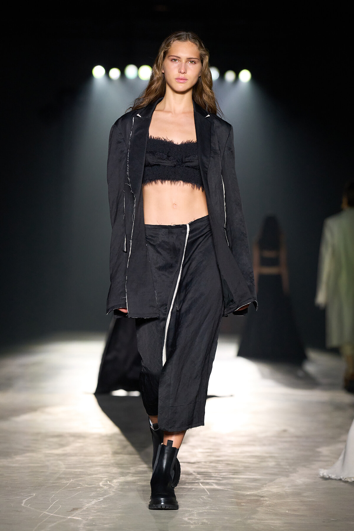 Model walks a fashion runway in an all-black outfit: cropped fringed top, oversized jacket, and matching pants with boots.