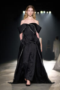 Model walks the runway in a full-length black off-the-shoulder gown with sculpted ruffles and white piping under dramatic backstage lighting.