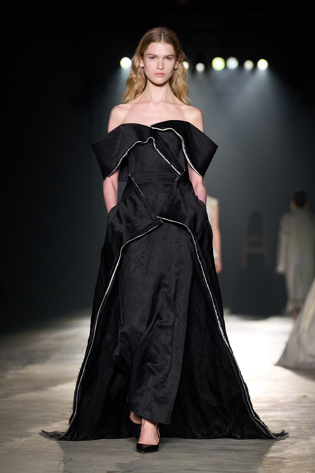 Model walks the runway in a full-length black off-the-shoulder gown with sculpted ruffles and white piping under dramatic backstage lighting.