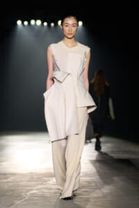 Model walks a dimly lit runway in an off-white sculptural sleeveless jumpsuit with irregular layered panels and fringed edges, spotlight behind.