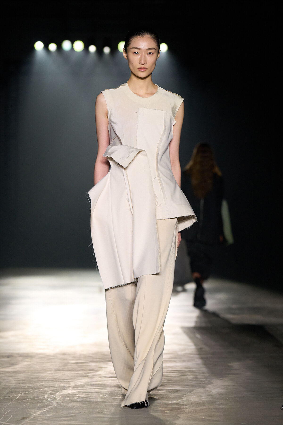 Model walks a dimly lit runway in an off-white sculptural sleeveless jumpsuit with irregular layered panels and fringed edges, spotlight behind.