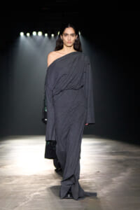 Female model on a runway wearing a loose, off-shoulder dark grey draped gown under bright stage lights.