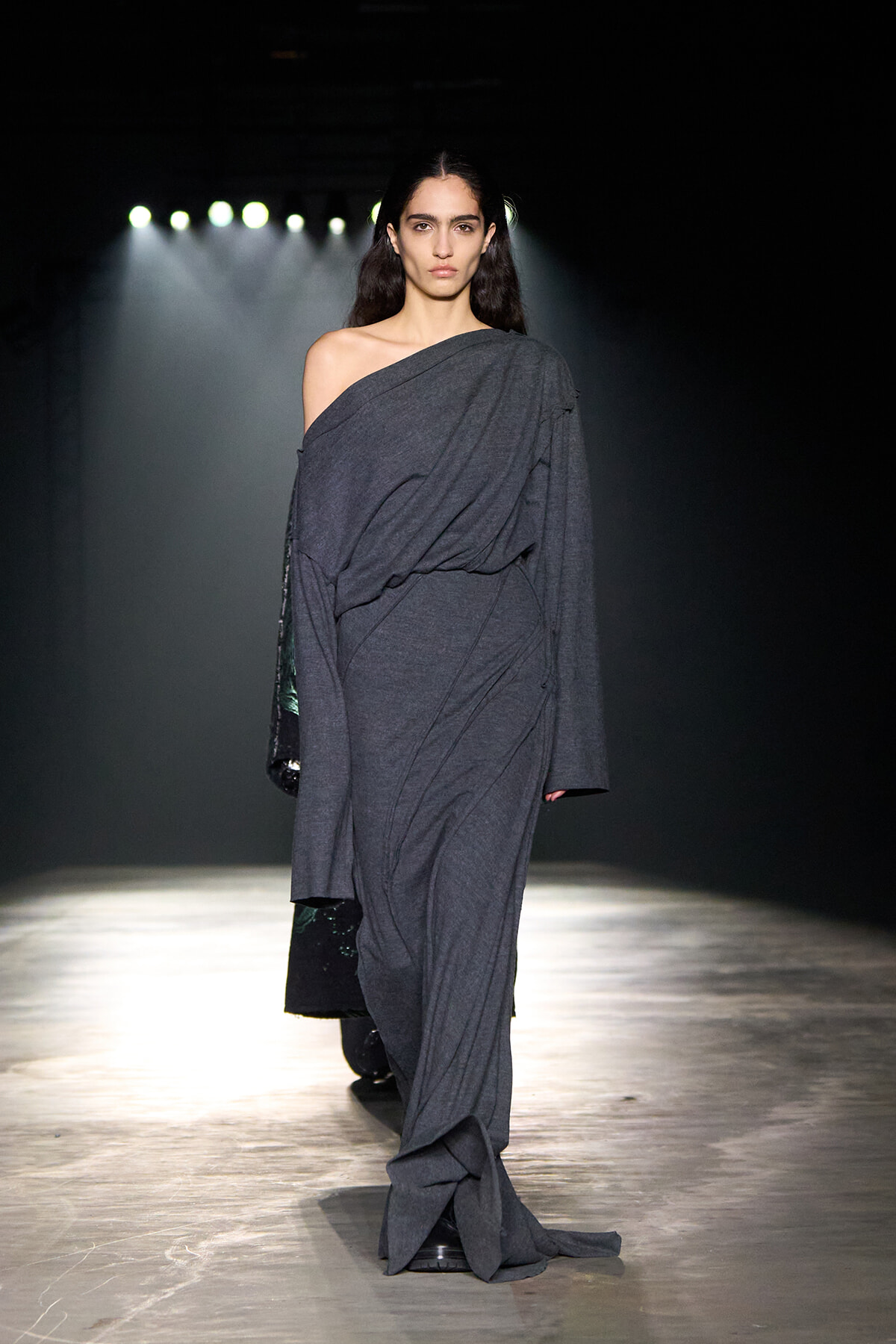 Female model on a runway wearing a loose, off-shoulder dark grey draped gown under bright stage lights.