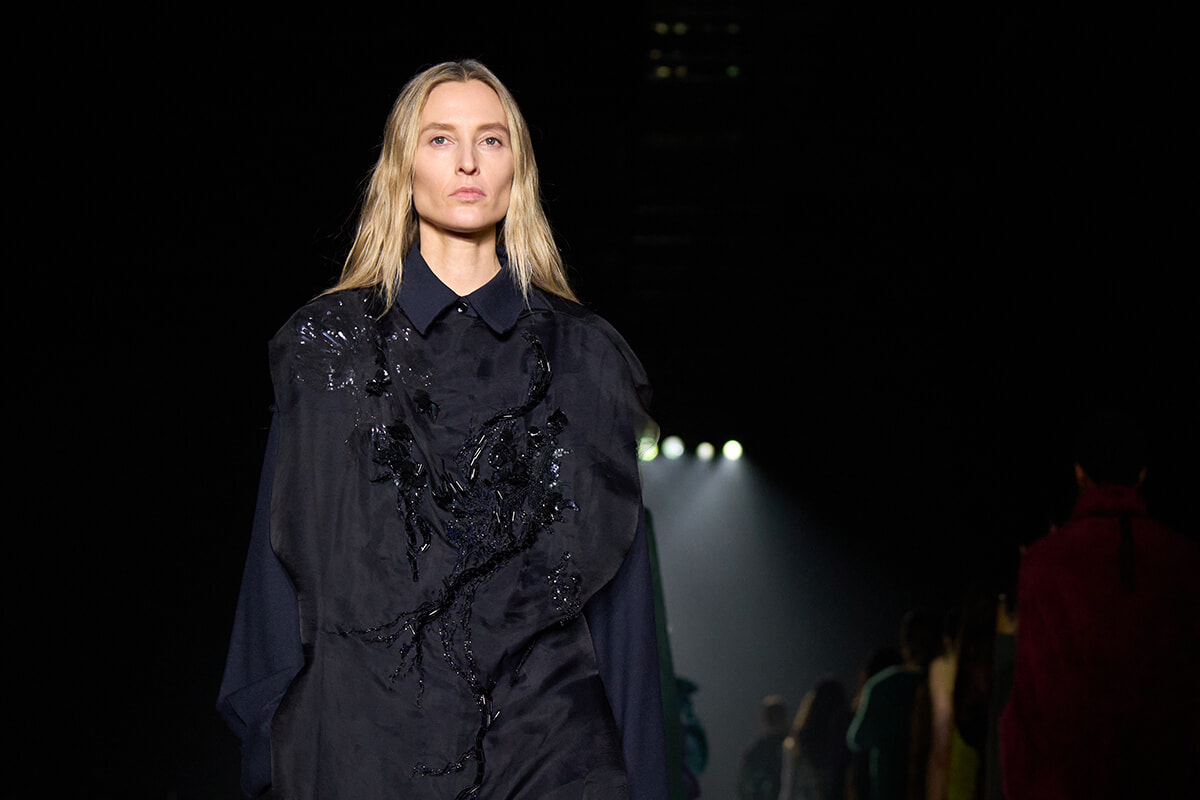 Fashion model walking a dark runway in a loose black outfit with sequined branch embroidery on the front and shoulders under bright spotlights.