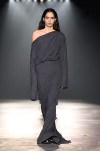 Model walking a runway in a dark gray, draped one-shoulder gown with long sleeves.