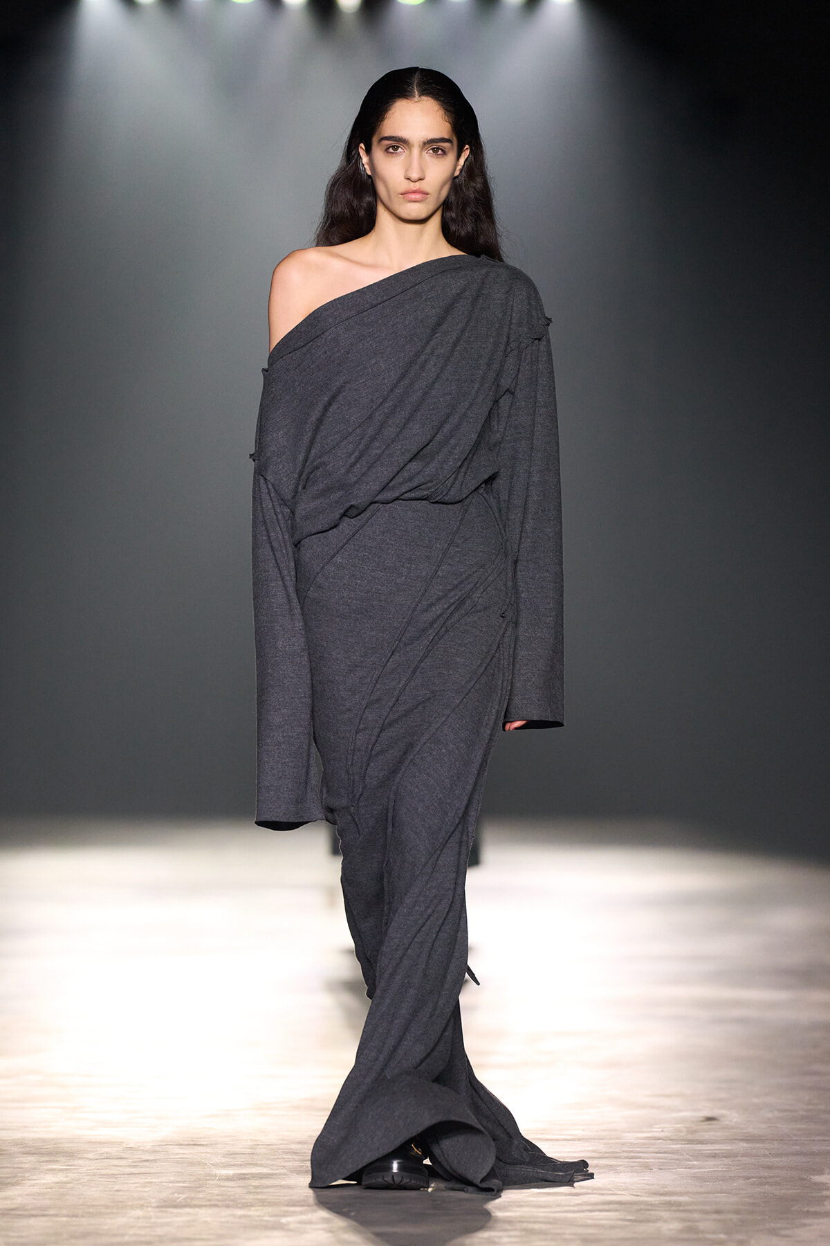 Model walking a runway in a dark gray, draped one-shoulder gown with long sleeves.