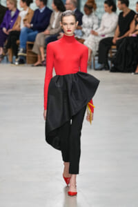 Model walks a fashion runway in a bright red turtleneck top and sculpted black draped skirt with red heels; audience blurred in background