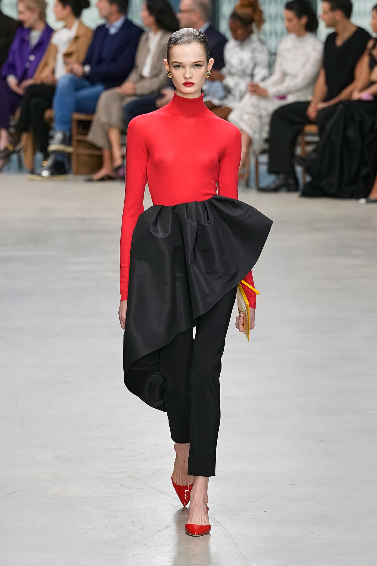 Model walks a fashion runway in a bright red turtleneck top and sculpted black draped skirt with red heels; audience blurred in background