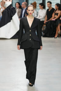 Model walks the runway in a black tailored blazer with gold buttons and peplum details, paired with matching black pants, audience in the background.