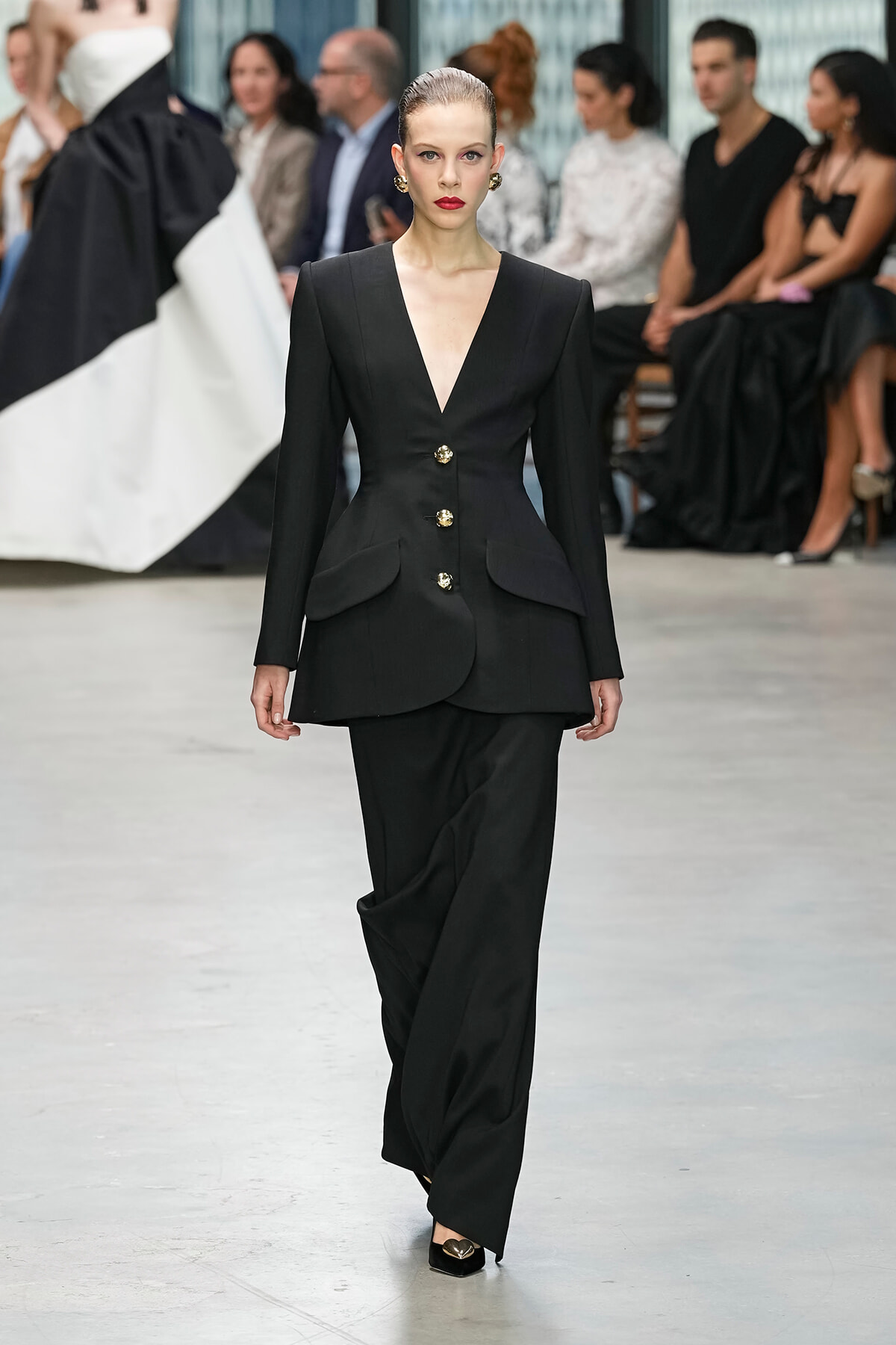 Model walks the runway in a black tailored blazer with gold buttons and peplum details, paired with matching black pants, audience in the background.