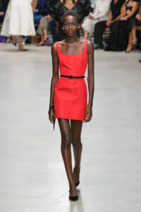 Model walks a runway wearing a red sleeveless dress with a slim black belt and black heels, audience out of focus in the background, fashion show setting.