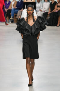 Model on a runway wearing a black outfit with an oversized, ruffled top and knee-length skirt/belt combo.