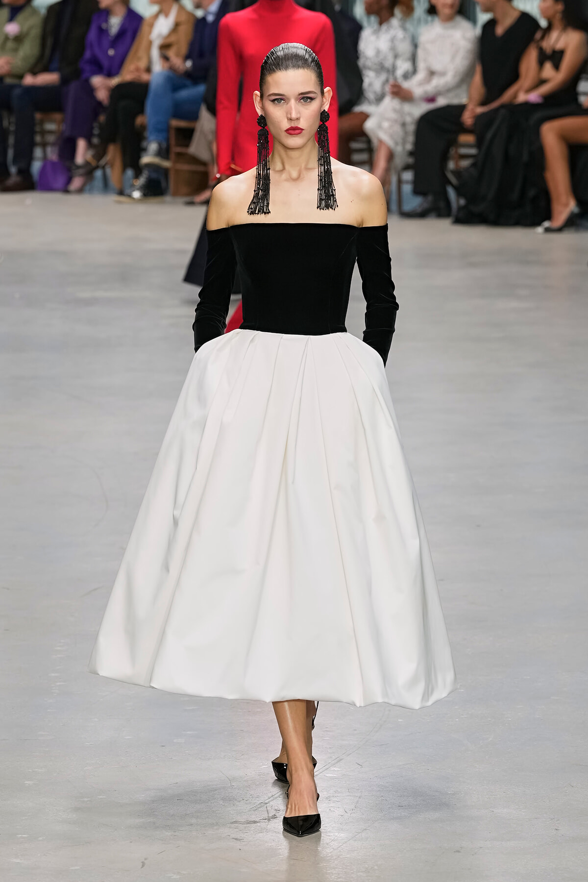 Model walking runway in off-shoulder black velvet top and full white skirt, black tassel earrings, red lipstick, focused expression