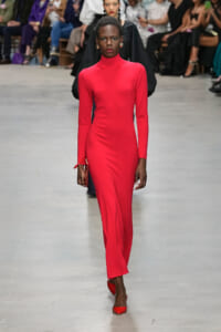Model walking a bright red, long-sleeve turtleneck dress down a fashion runway with spectators in the background.