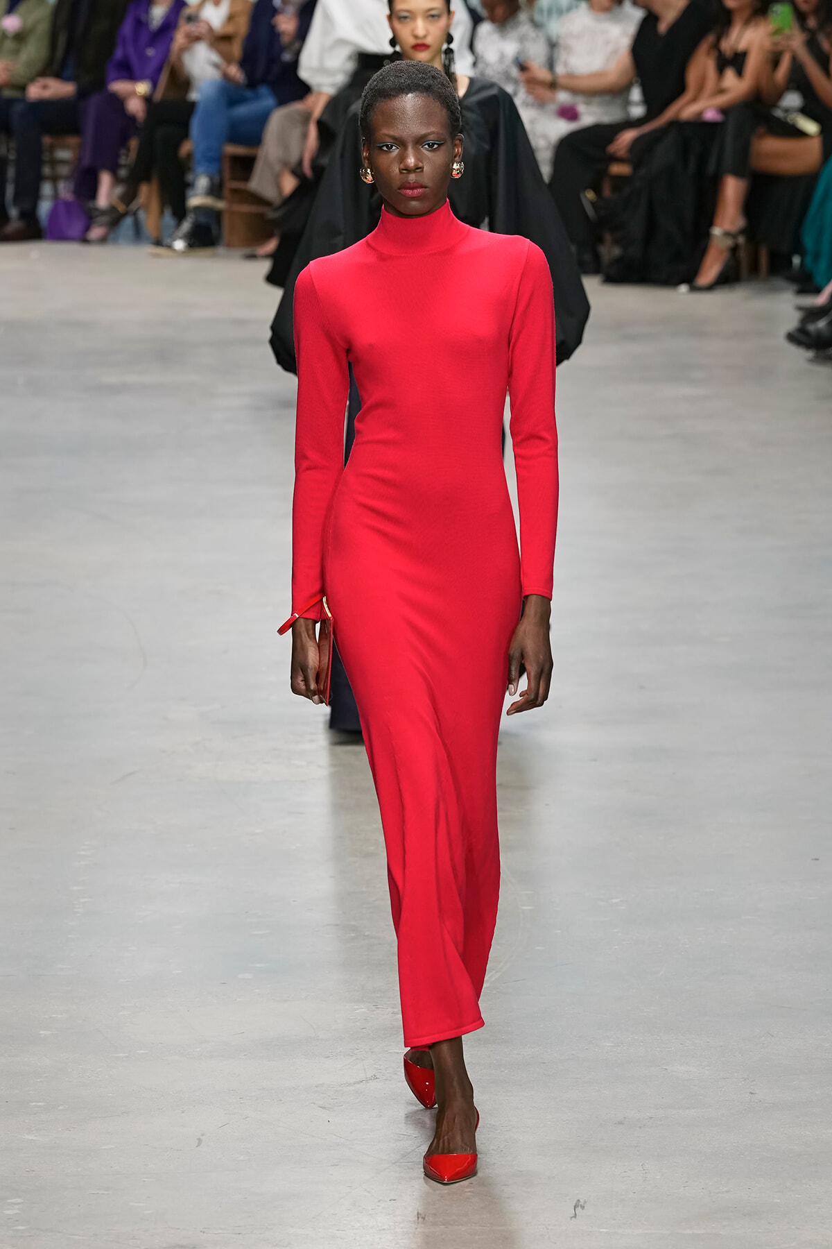 Model walking a bright red, long-sleeve turtleneck dress down a fashion runway with spectators in the background.
