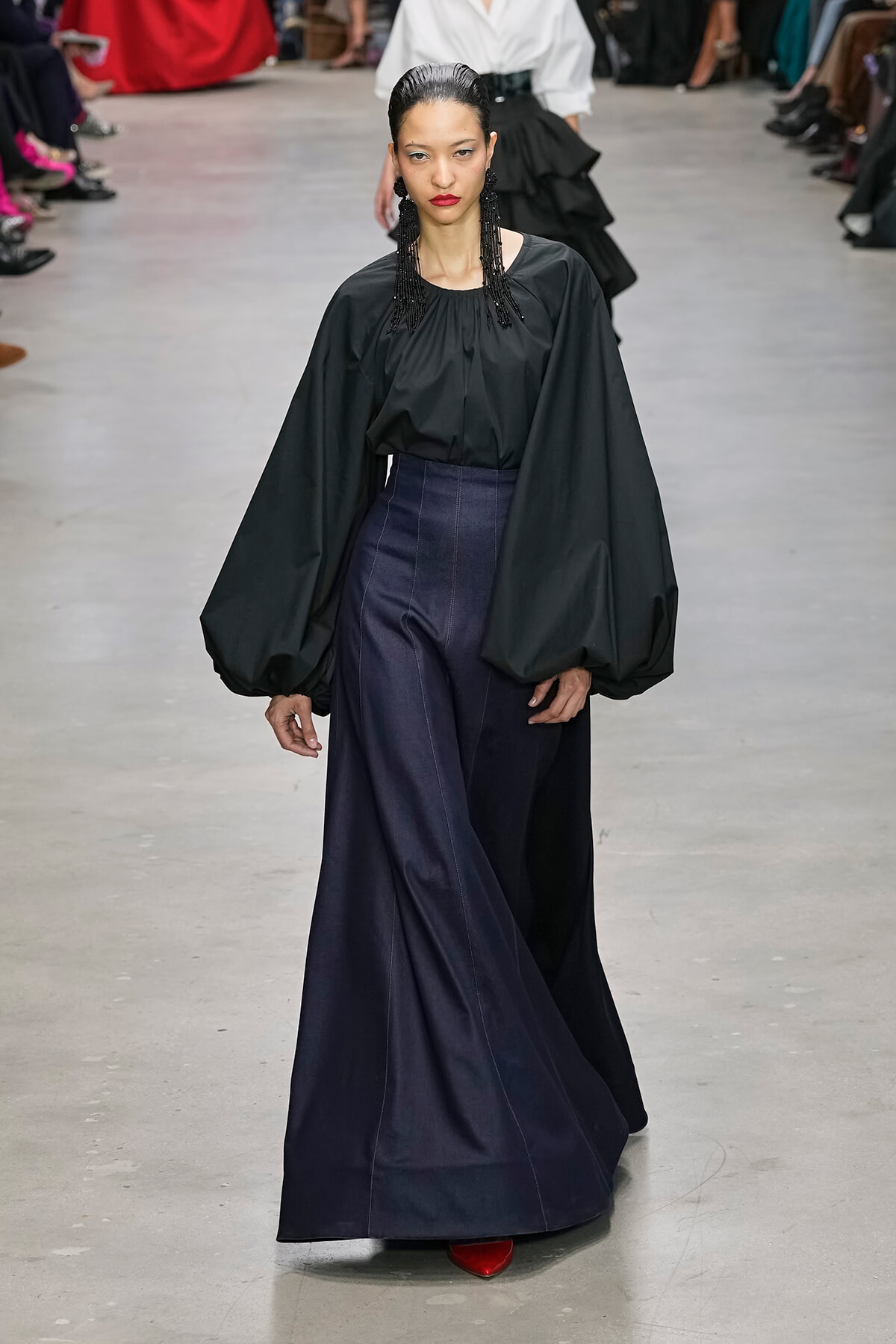 Model walking a fashion runway in a black voluminous-sleeve blouse and navy wide-leg skirt, red shoes peeking from beneath.