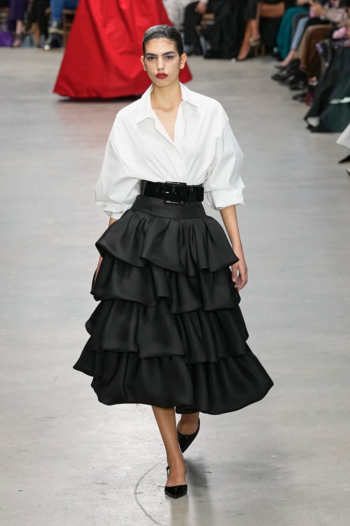 Runway model in a white button-up blouse with oversized sleeves and a voluminous black tiered skirt with a wide belt.