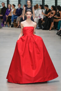Model in a bright red strapless ball gown walking the runway with a seated audience in the background, watching.