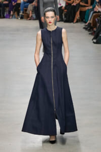 Model walks a full-length navy sleeveless gown with a gold front zipper on a runway, audience blurred in the background.