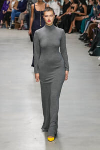 Model walks a fashion runway in a fitted gray turtleneck dress, with yellow shoes and a blurred audience in the background.