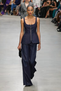 Model walks down runway in a navy sleeveless jumpsuit with front zipper and curved peplum, accessorized with gold hoop earrings.