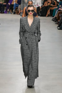 Model walks a black-and-white checkered suit with belt, wearing dark sunglasses on a runway. Audience blurred in background.