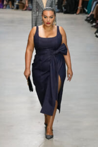 Model walks a runway in a navy sleeveless midi dress with a waist-tie bow and a high slit, accessorized with statement earrings and checked heels.