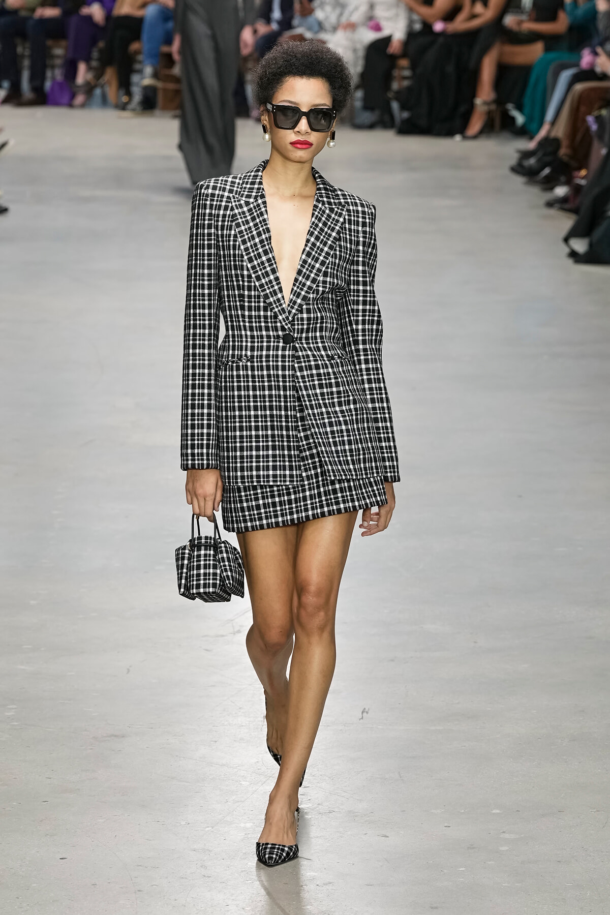 Model walks the runway in a black-and-white plaid blazer and matching mini skirt, carrying a small plaid handbag and wearing oversized sunglasses.