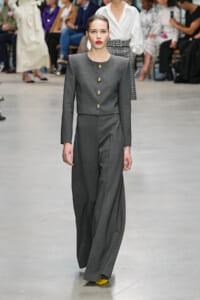 Model wearing a gray tailored pant suit with gold buttons on a runway, pairing with bright yellow shoes as audience watches in the background.