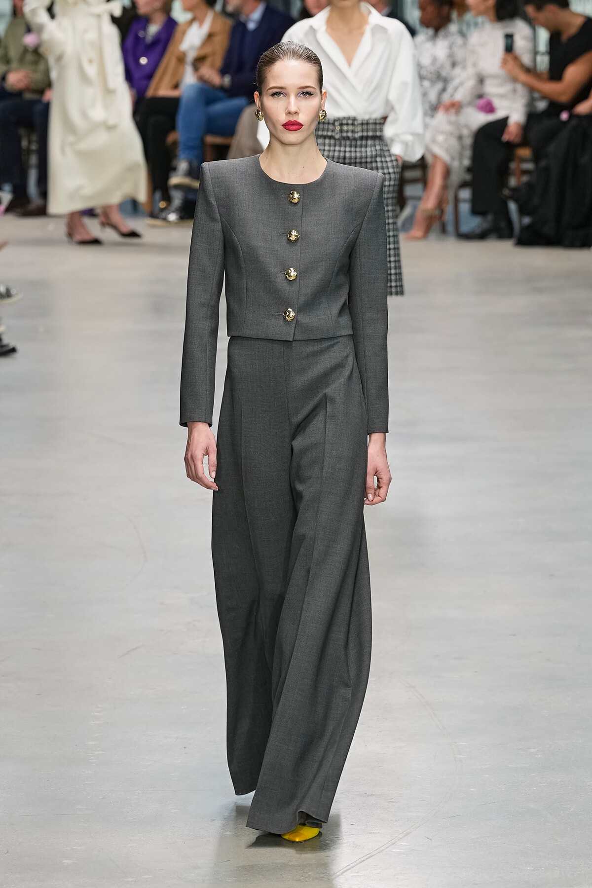 Model wearing a gray tailored pant suit with gold buttons on a runway, pairing with bright yellow shoes as audience watches in the background.